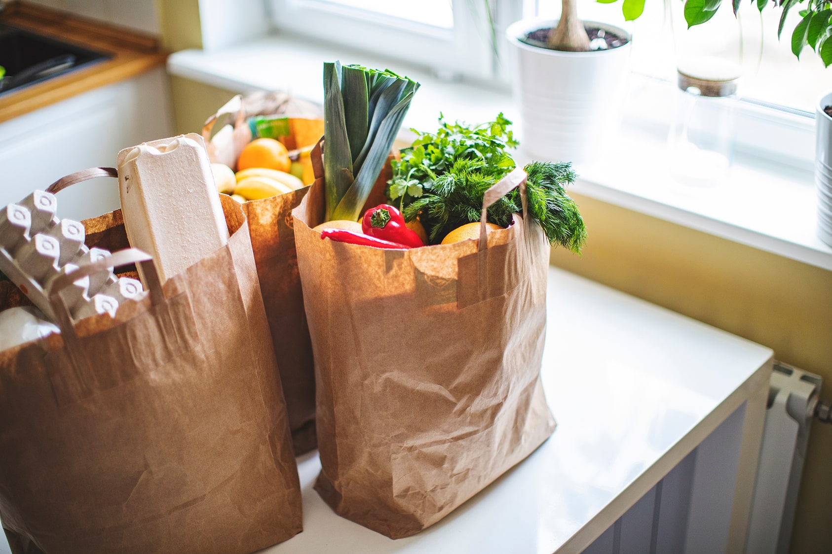 Image of groceries in brown paper bags on the table next to a bright window.